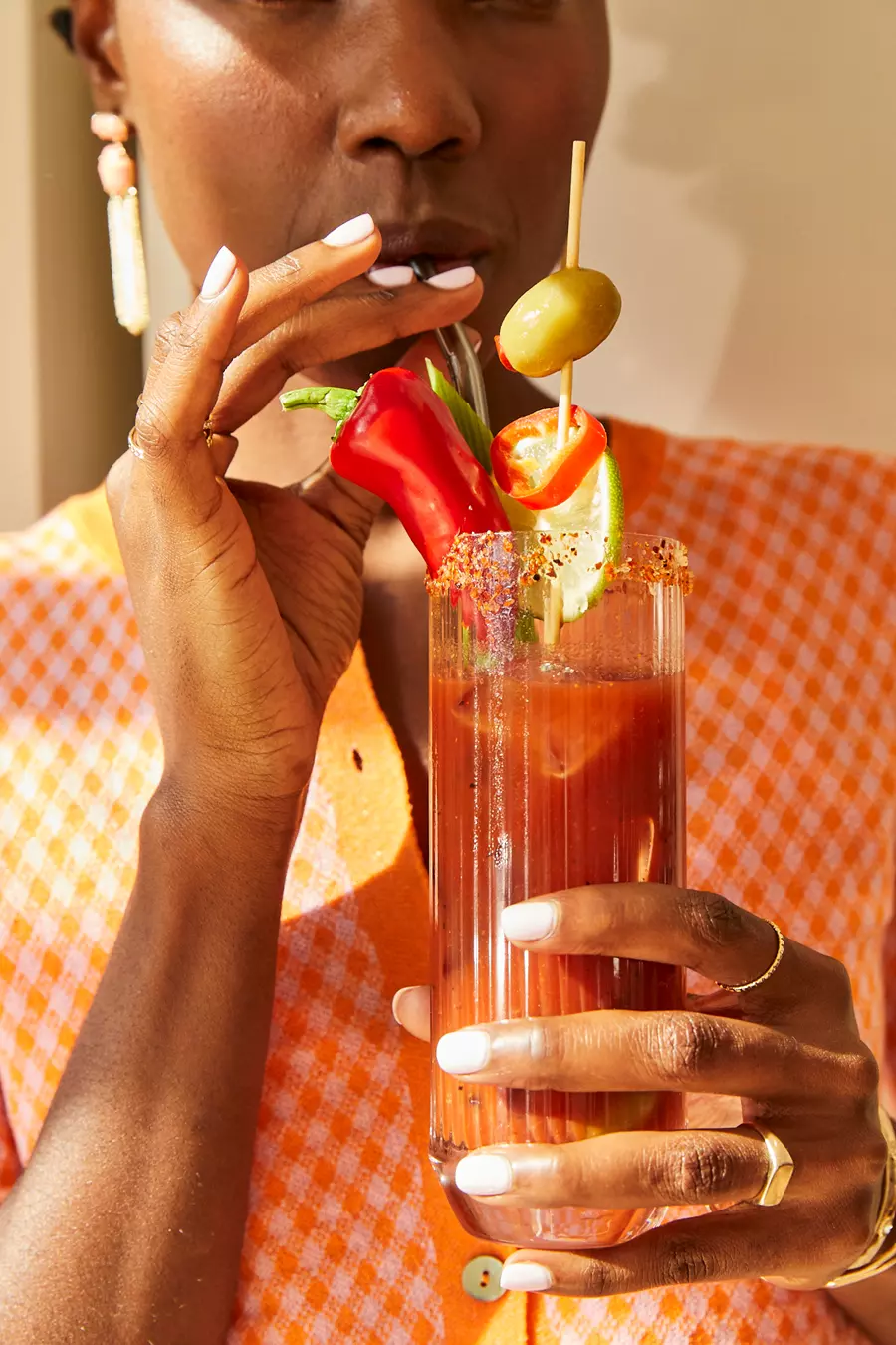 A person holding a Bloody Mary cocktail garnished with an olive, peppers, and celery, wearing an orange checkered shirt and gold jewelry.