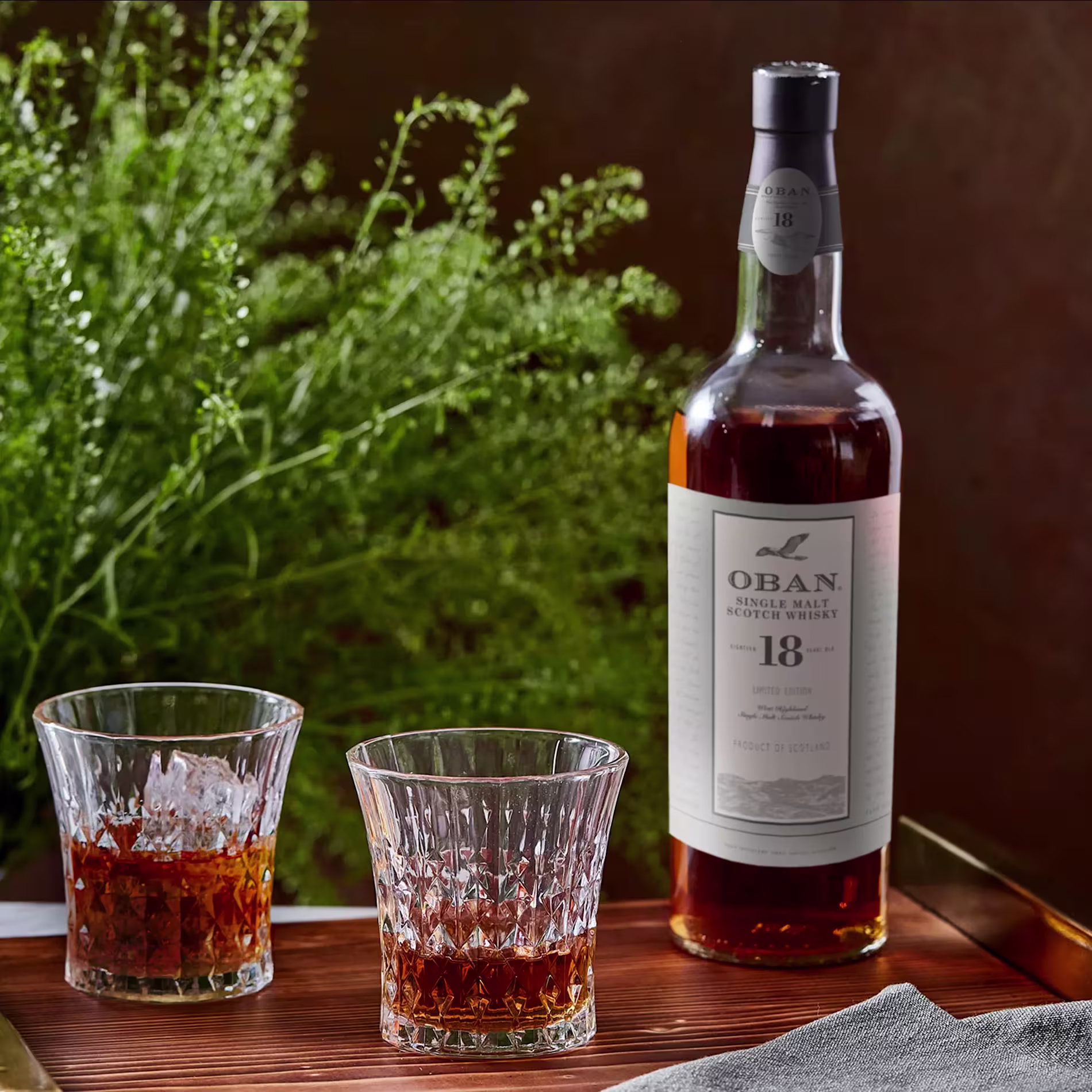 A bottle of Oban 18 Year Old Single Malt Scotch Whisky stands beside two filled glasses on a tray, with green foliage in the background.