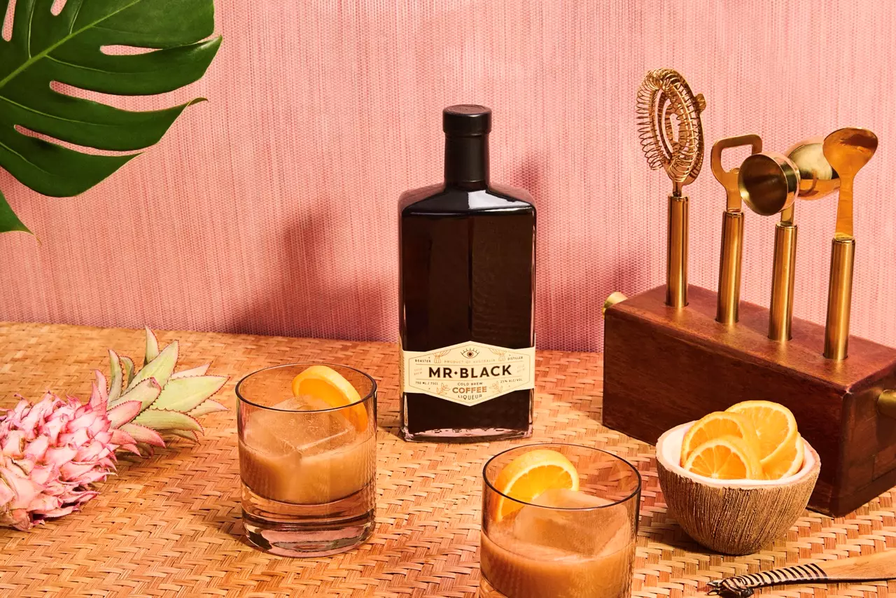A bottle of Mr. Black Coffee Liqueur, two glasses with iced drinks and orange slices, a cocktail tool set, sliced oranges, and a pink pineapple on a woven surface with a pink background.