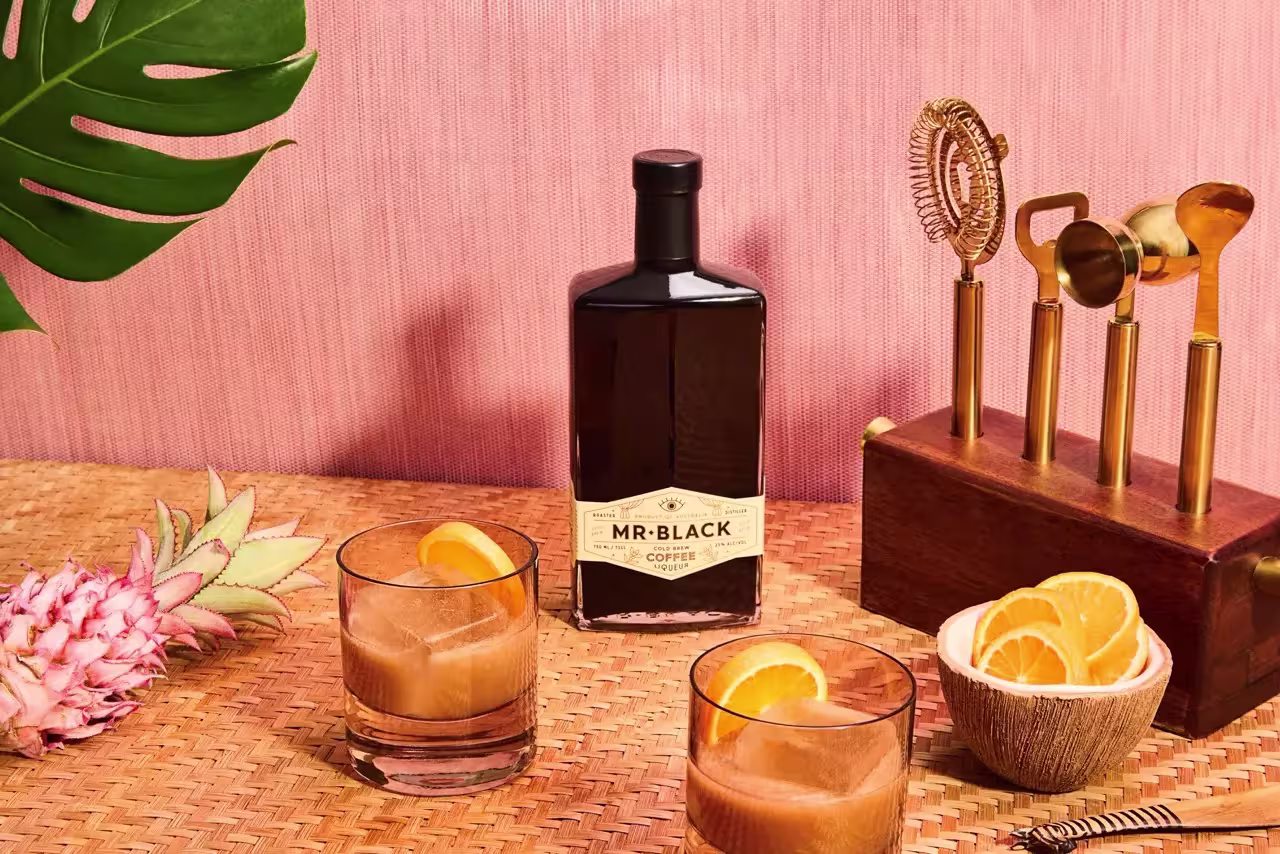 A bottle of Mr. Black Coffee Liqueur, two glasses with iced drinks and orange slices, a cocktail tool set, sliced oranges, and a pink pineapple on a woven surface with a pink background.