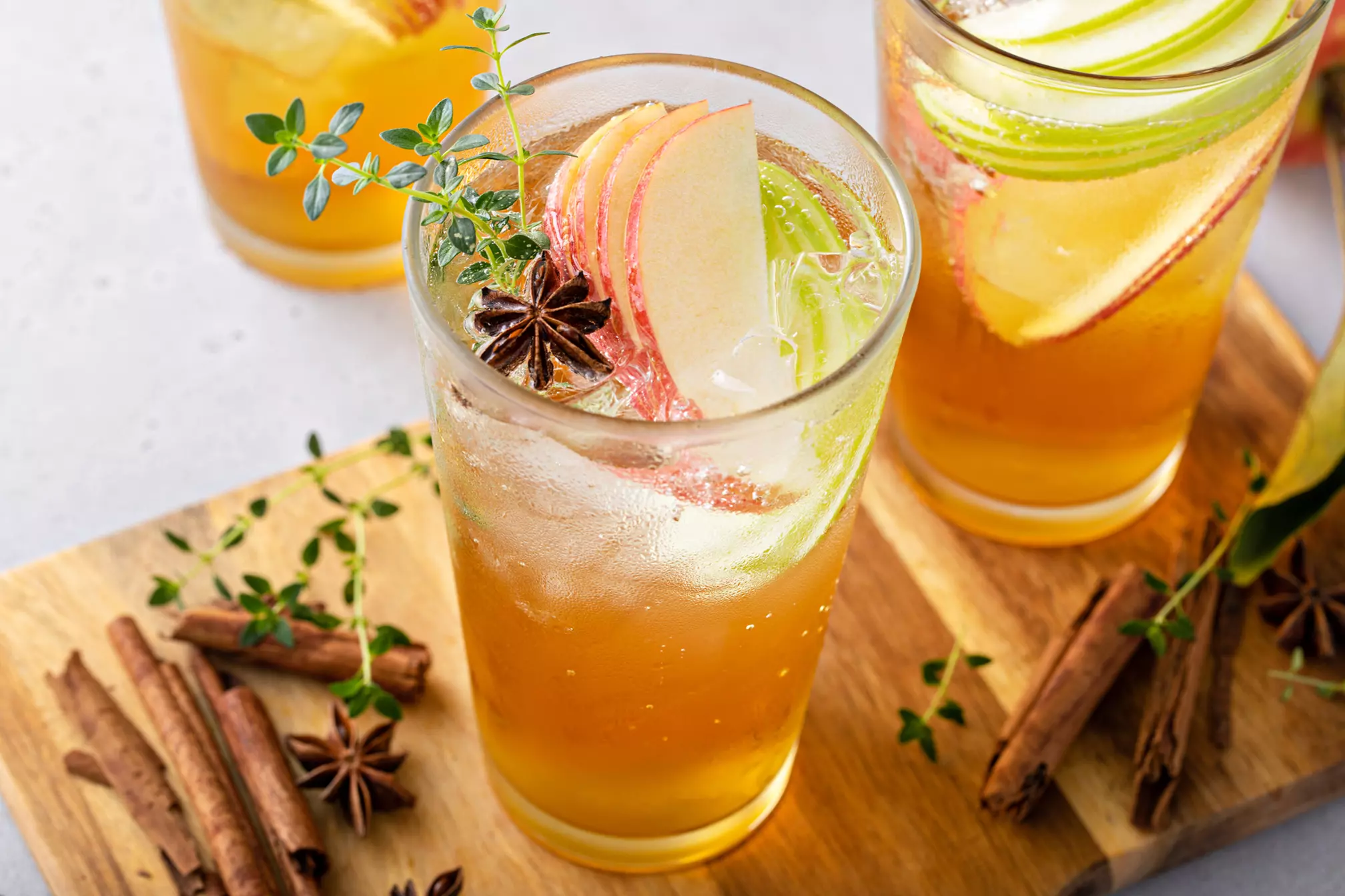 Iced apple cider in a tall glass garnished with apple slices, star anise, thyme, and cinnamon sticks on a wooden board.