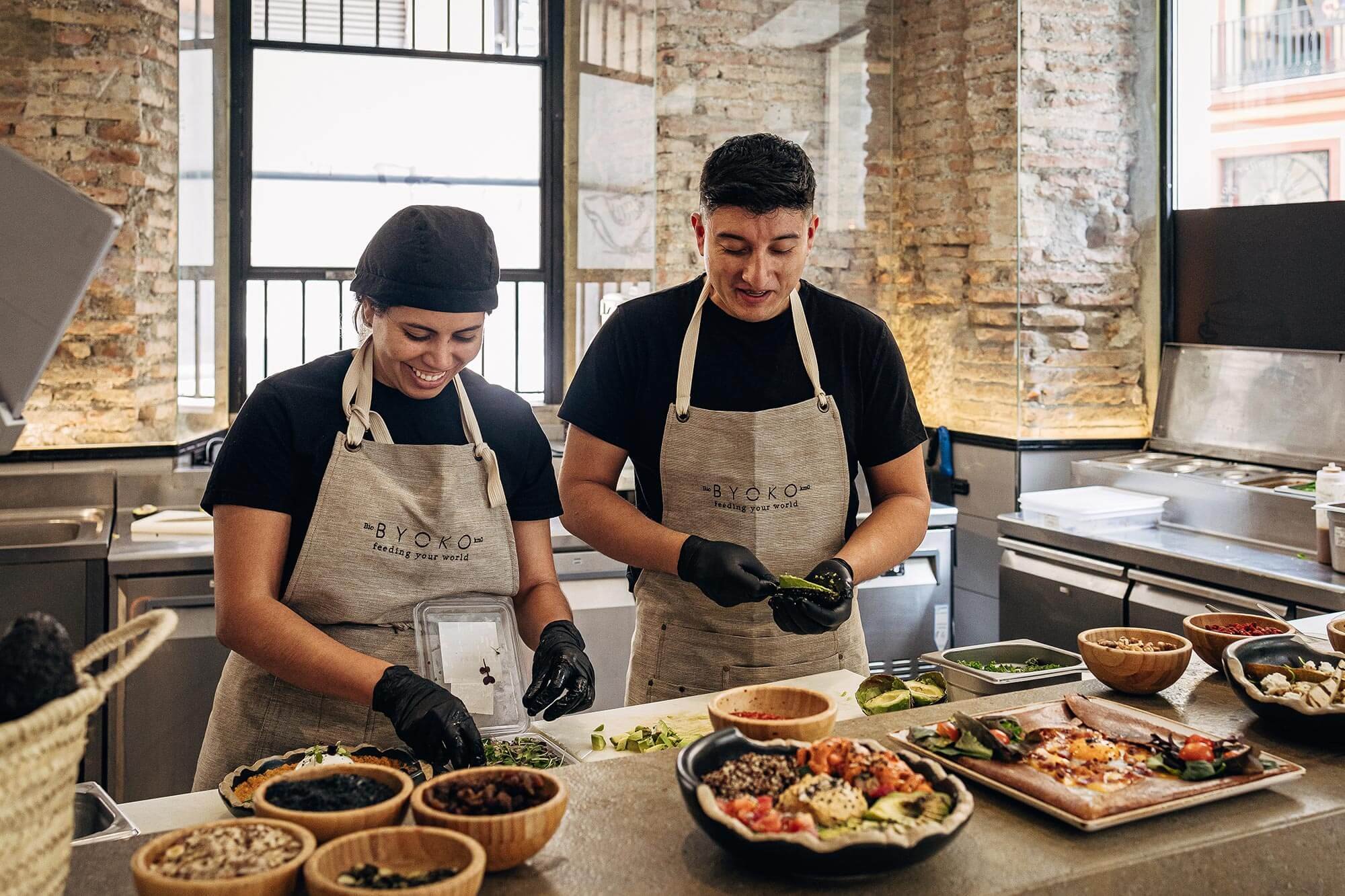 Cocina de Byoko con dos personas preparando comida, rodeadas de ingredientes frescos y platos variados sobre una mesa en un entorno moderno.