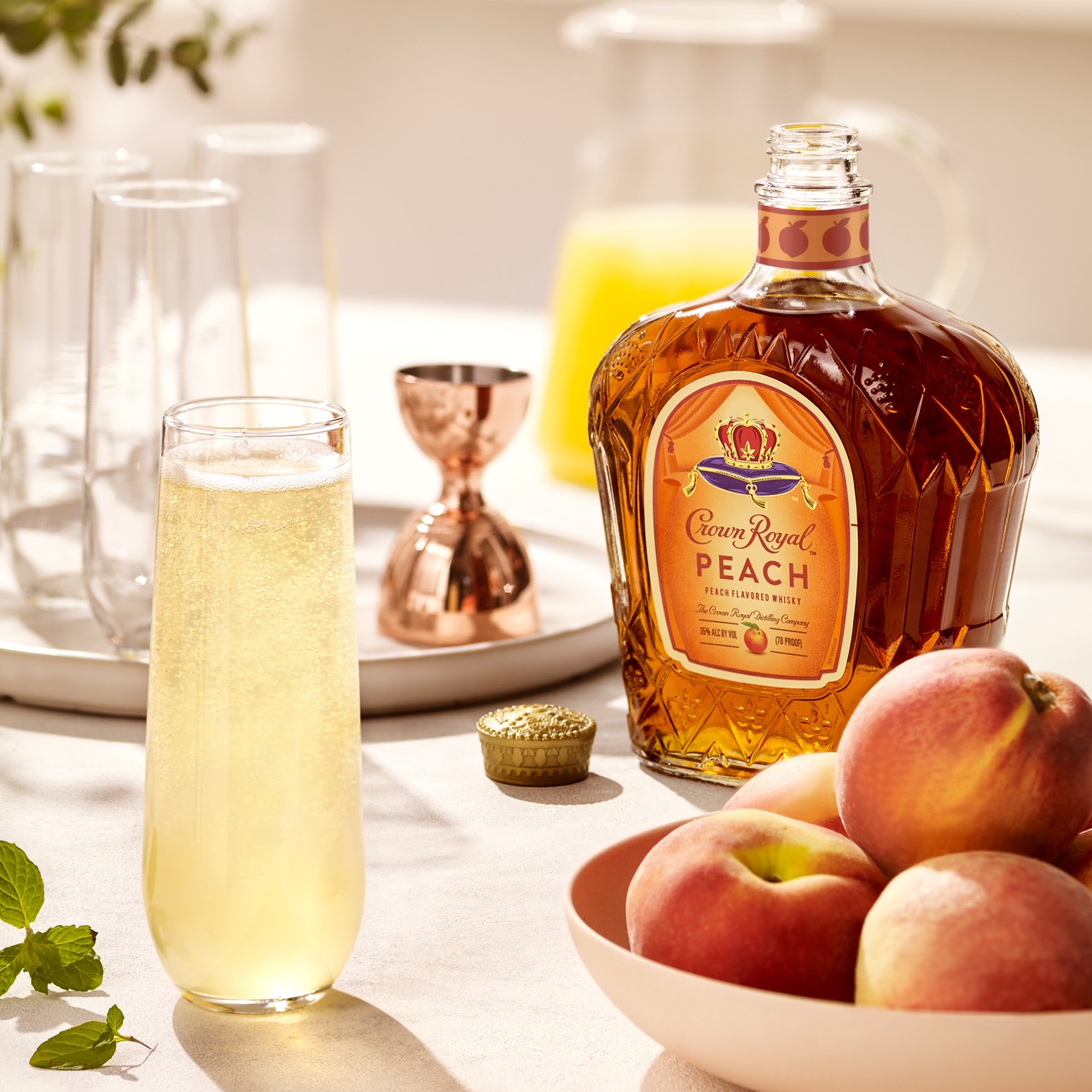 A bottle of Crown Royal Peach whisky sits next to a glass of yellow cocktail, a bowl of peaches, a jigger, and a pitcher of juice on a sunlit table.
