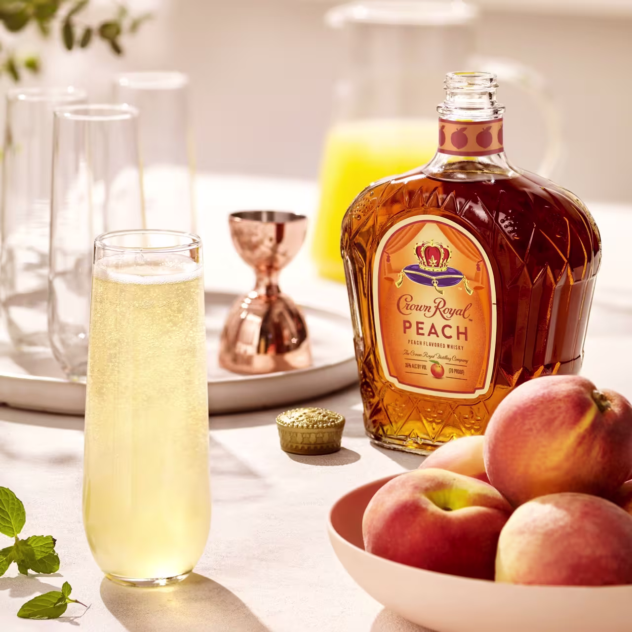 A bottle of Crown Royal Peach whisky sits next to a glass of yellow cocktail, a bowl of peaches, a jigger, and a pitcher of juice on a sunlit table.