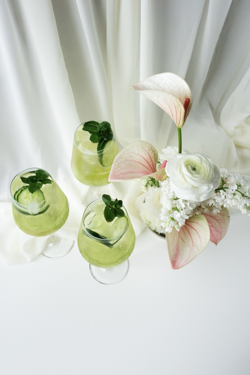 Three green drinks garnished with mint and cucumber sit next to a white floral arrangement with light pink anthuriums on a white surface with a white fabric backdrop.