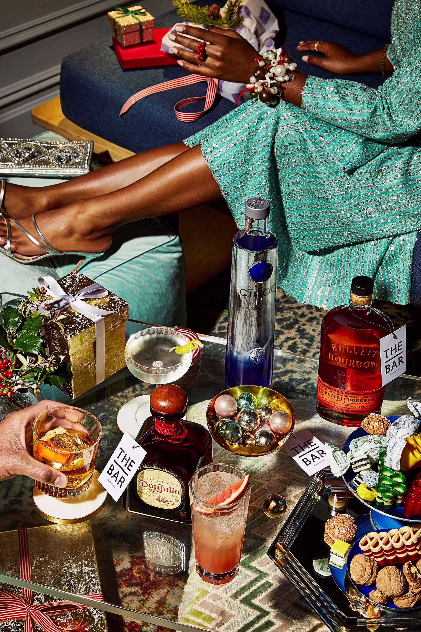 A festive table with assorted cocktails, spirits, snacks, wrapped gifts, and holiday decorations; people’s hands and legs are partially visible.
