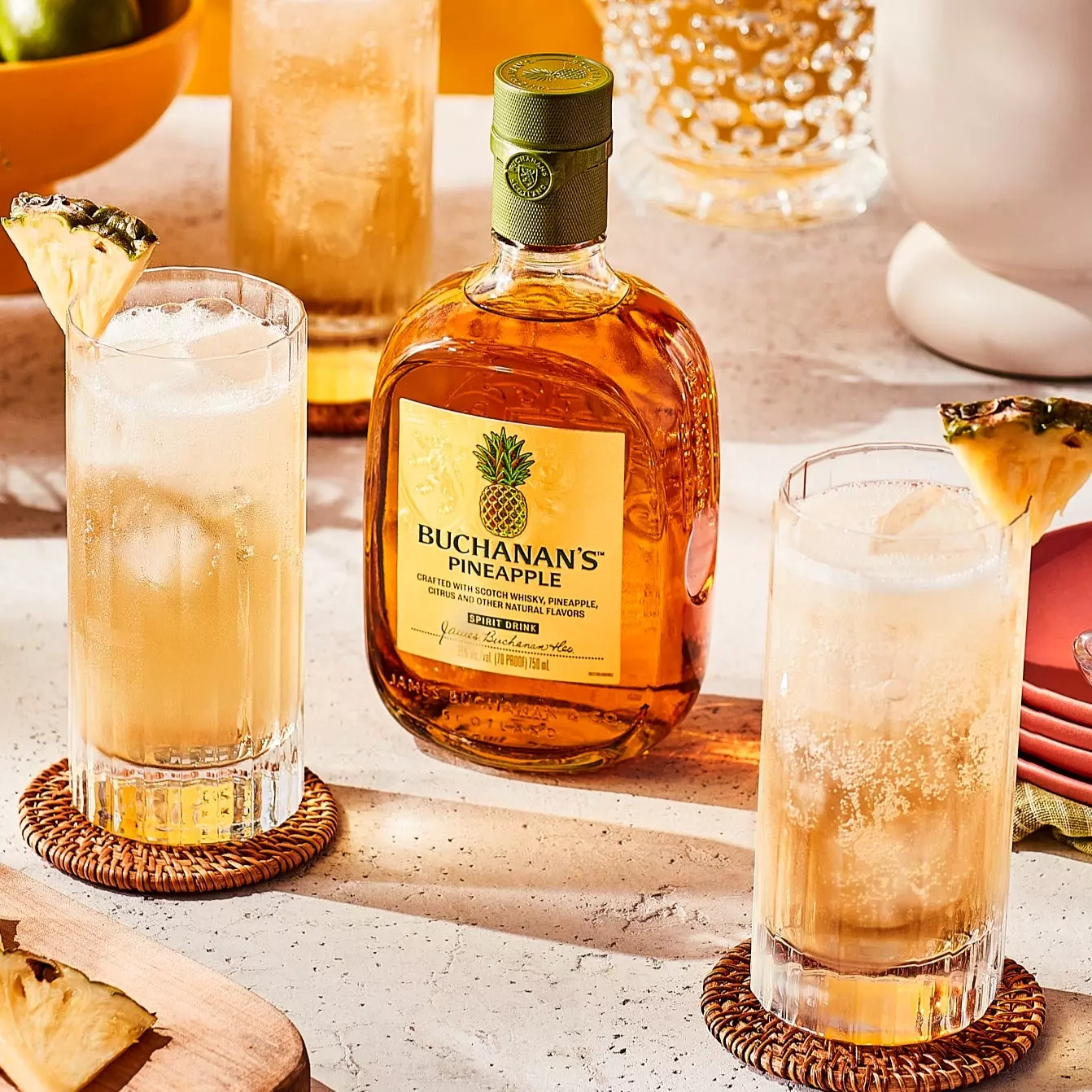 A bottle of Buchanan’s Pineapple whisky is placed on a table with two cocktails garnished with pineapple slices.