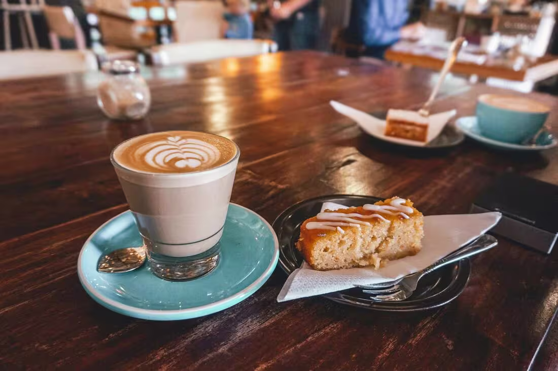 A glass of latte with latte art sits on a blue saucer beside a plate with a slice of iced cake on a wooden table in a café.