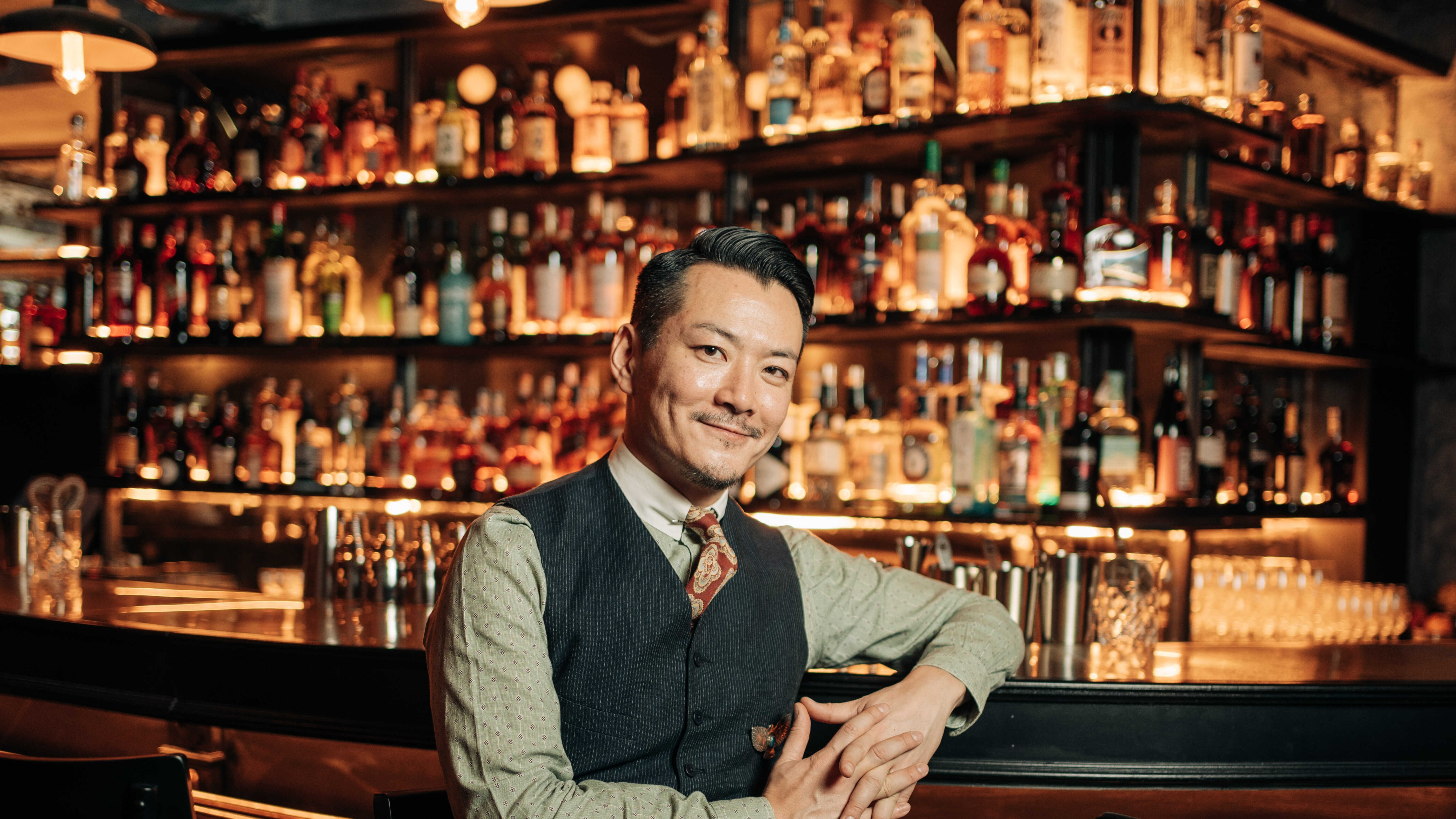 Bartender posando con elegancia frente a una barra repleta de botellas iluminadas en un ambiente cálido y sofisticado.