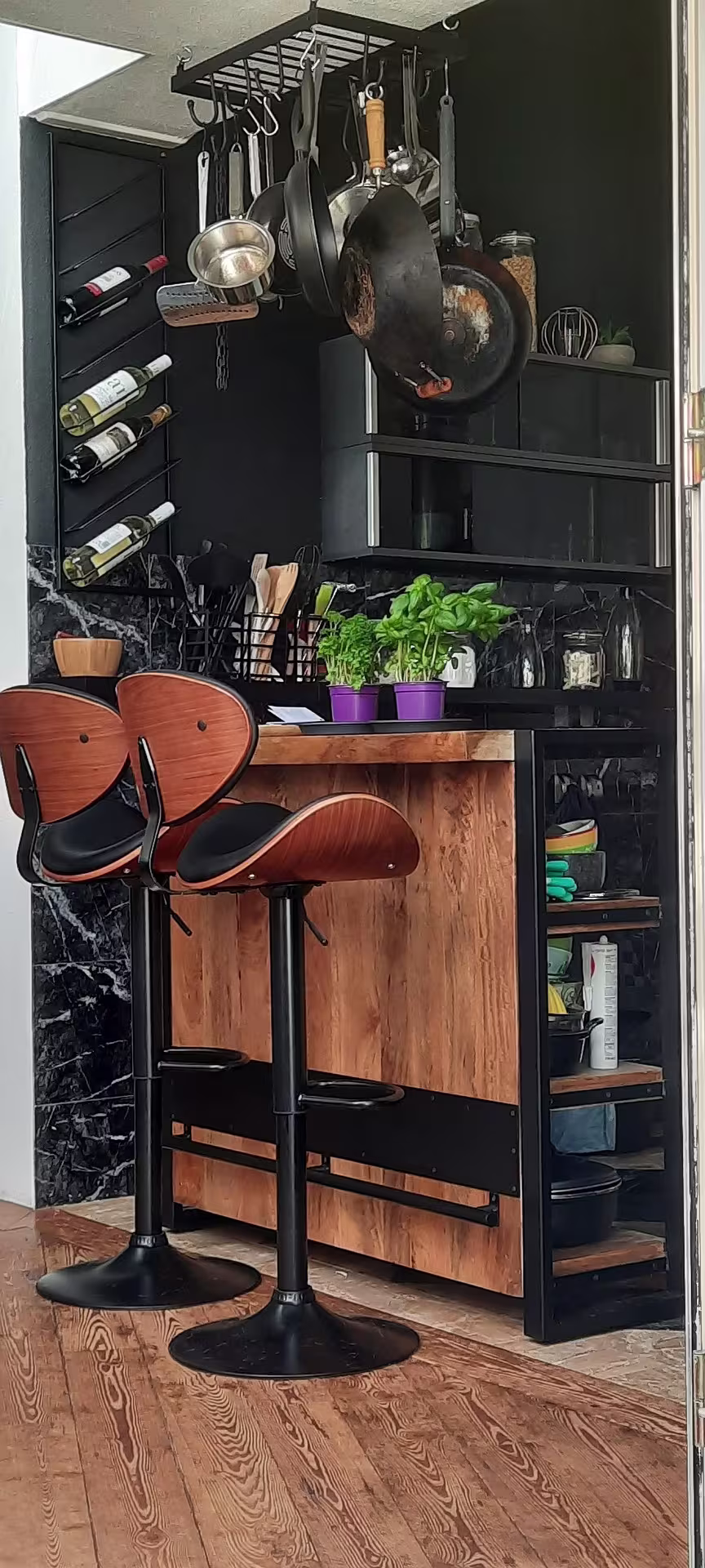 A compact kitchen with a wooden island, two bar stools, hanging pots and pans, a wine rack, potted herbs on the counter, and black shelves holding kitchen items.