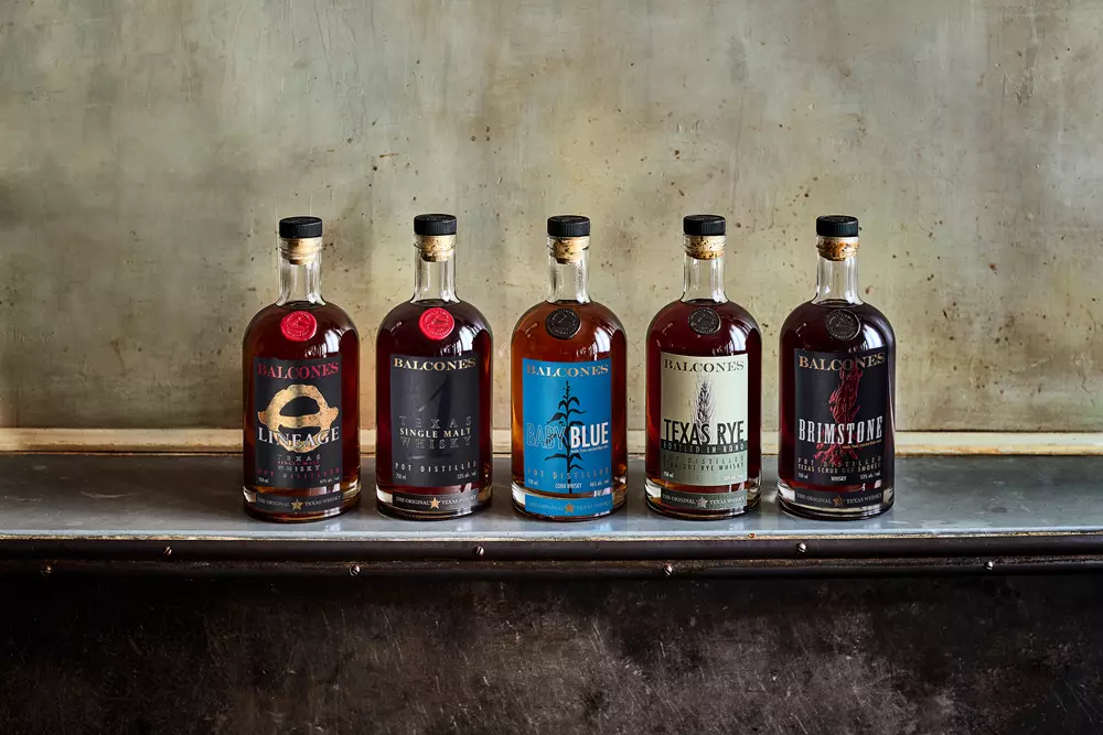 Five bottles of Balcones whiskey are lined up on a metal surface against a textured wall, each featuring distinct labels and colors.