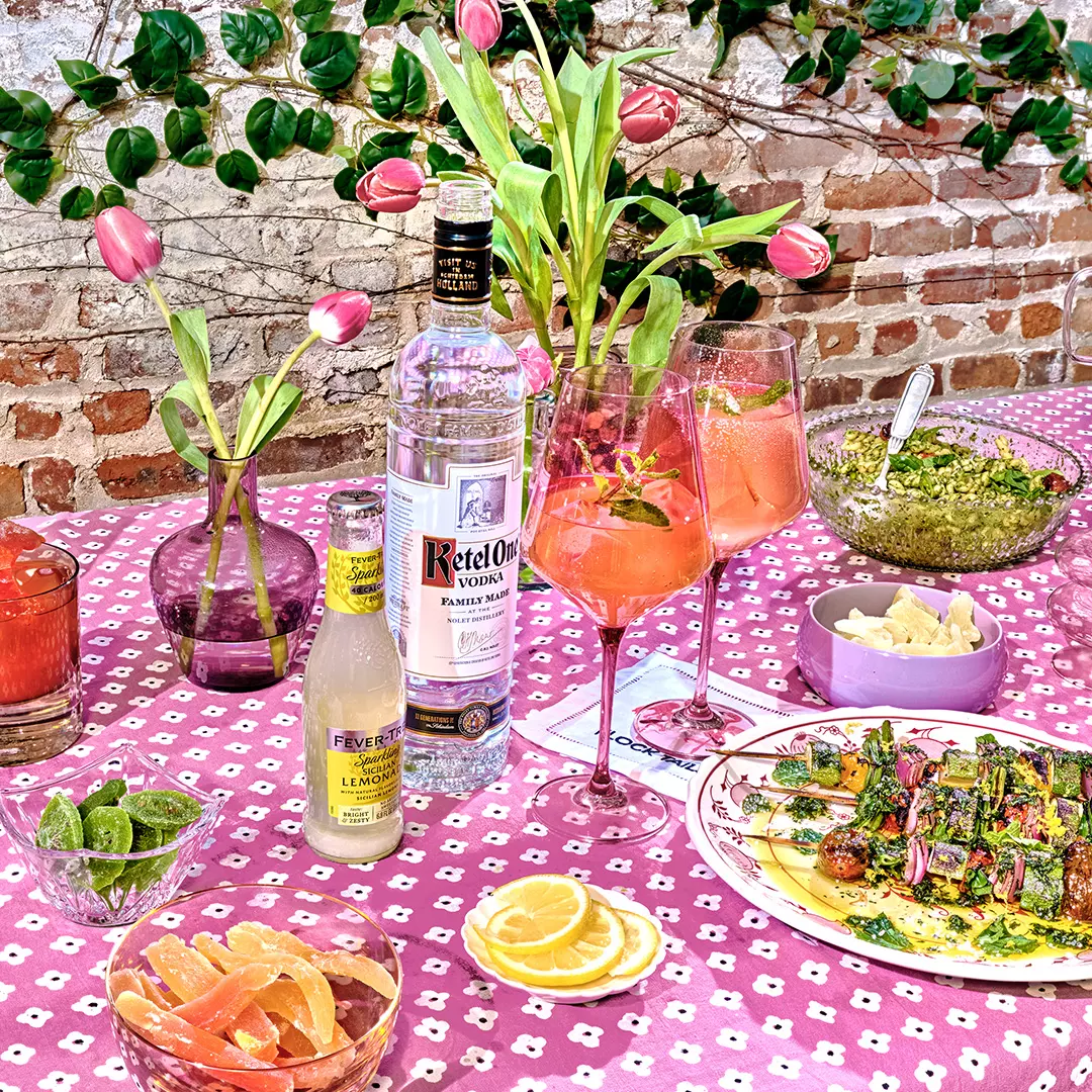 A pink-patterned table is set with vodka, tonic, cocktails, sliced citrus, mixed drinks, a salad, guacamole with chips, and vases of pink tulips, against a brick wall backdrop.