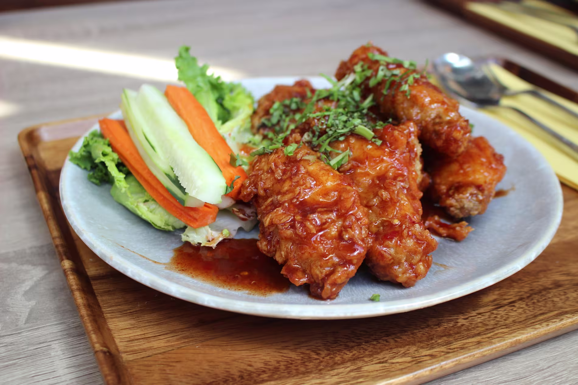 A plate of fried chicken wings coated in sauce, garnished with chopped herbs, served with lettuce, cucumber, and carrot sticks on a wooden tray.