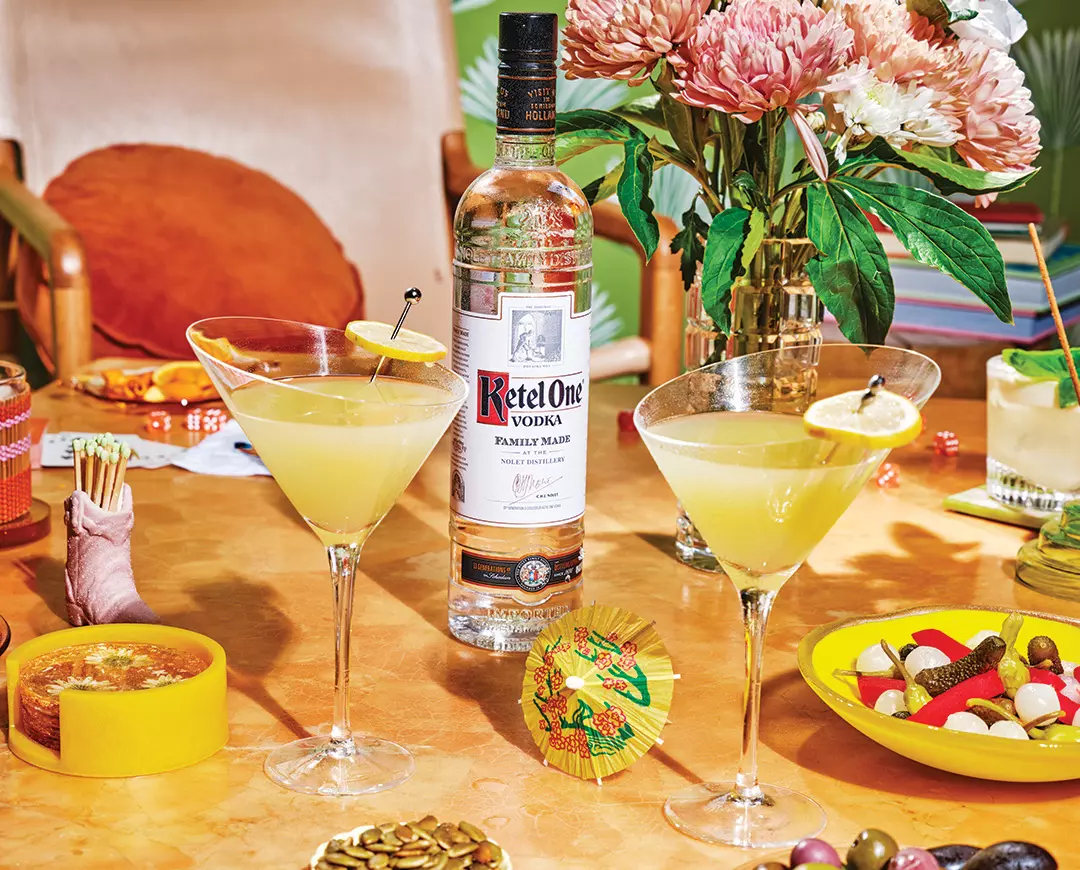 A bottle of Ketel One vodka is placed behind two martini glasses with yellow cocktails, surrounded by snacks, flowers, and assorted table items on an orange surface.