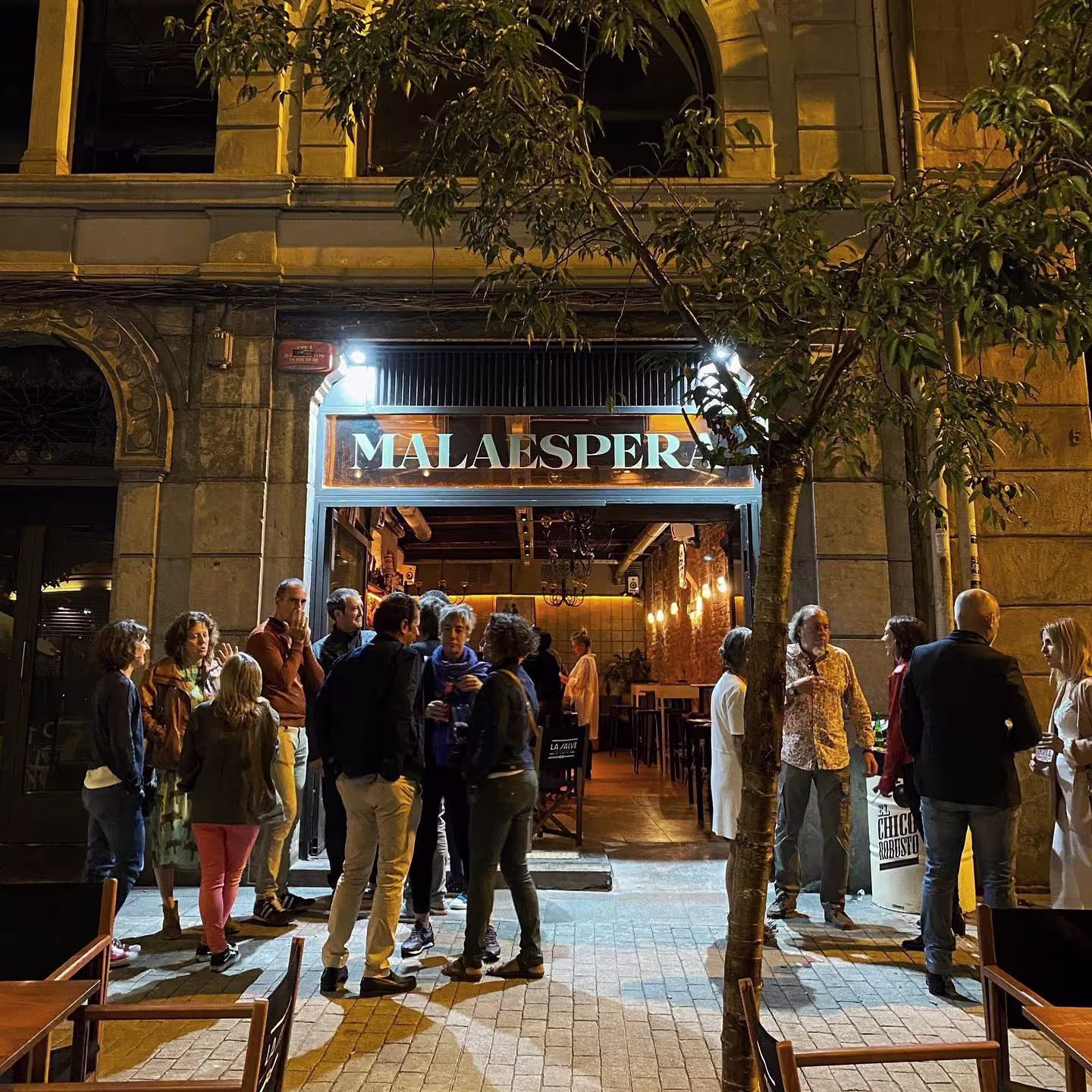 Fachada iluminada de Malaespera con grupos de personas conversando en la entrada, mobiliario de terraza y ambiente nocturno animado.