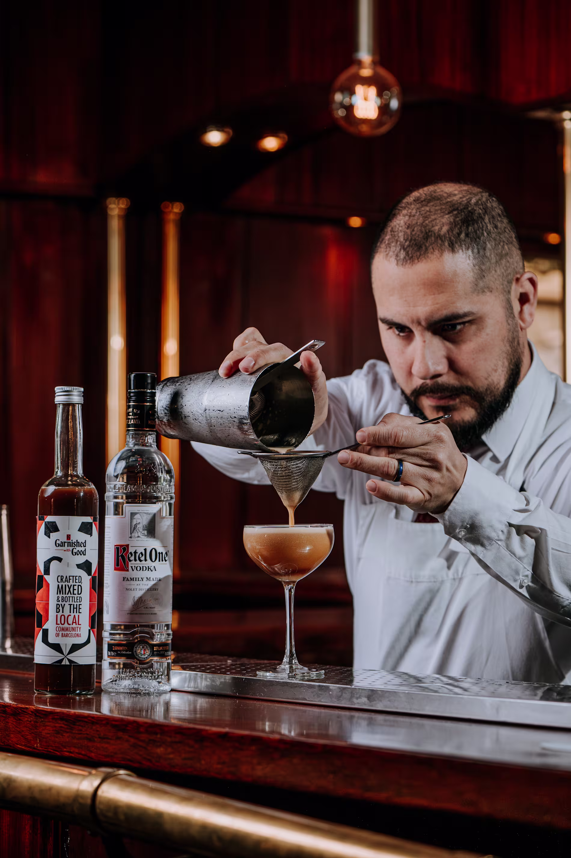 Bartender sirviendo un cóctel con doble colado en copa tipo cóctel. A su lado, botellas de Ketel One y Garnished with Good sobre una barra de madera elegante con luz cálida.