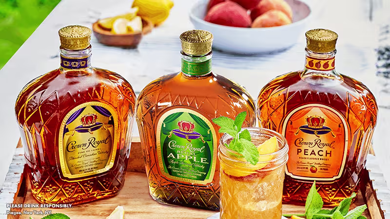 Three bottles of Crown Royal whiskey—original, apple, and peach flavors—sit on a table next to a cocktail garnished with mint and fruit slices, with peaches in the background.