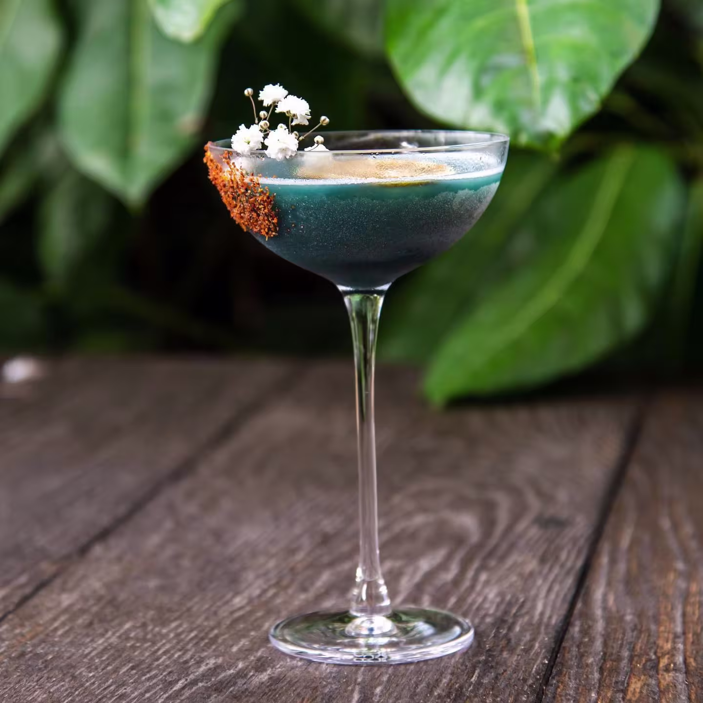 Cóctel azul oscuro en copa elegante, decorado con flores blancas y borde de sal especiada, sobre mesa de madera con hojas verdes de fondo.