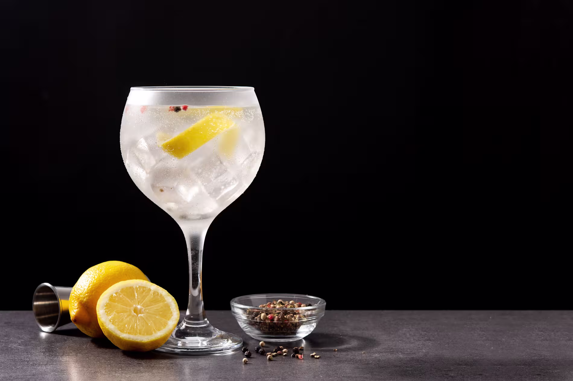 A gin and tonic with ice and a lemon slice in a balloon glass, next to a halved lemon, a jigger, and a small bowl of peppercorns on a dark surface.