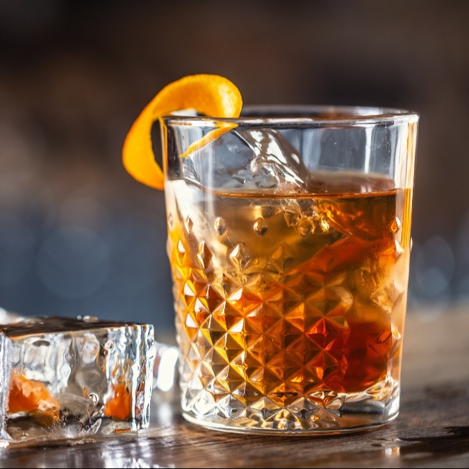 Un cóctel de ron en un vaso de whisky con un cubito grande de hielo y una tira de piel de naranja retorcida