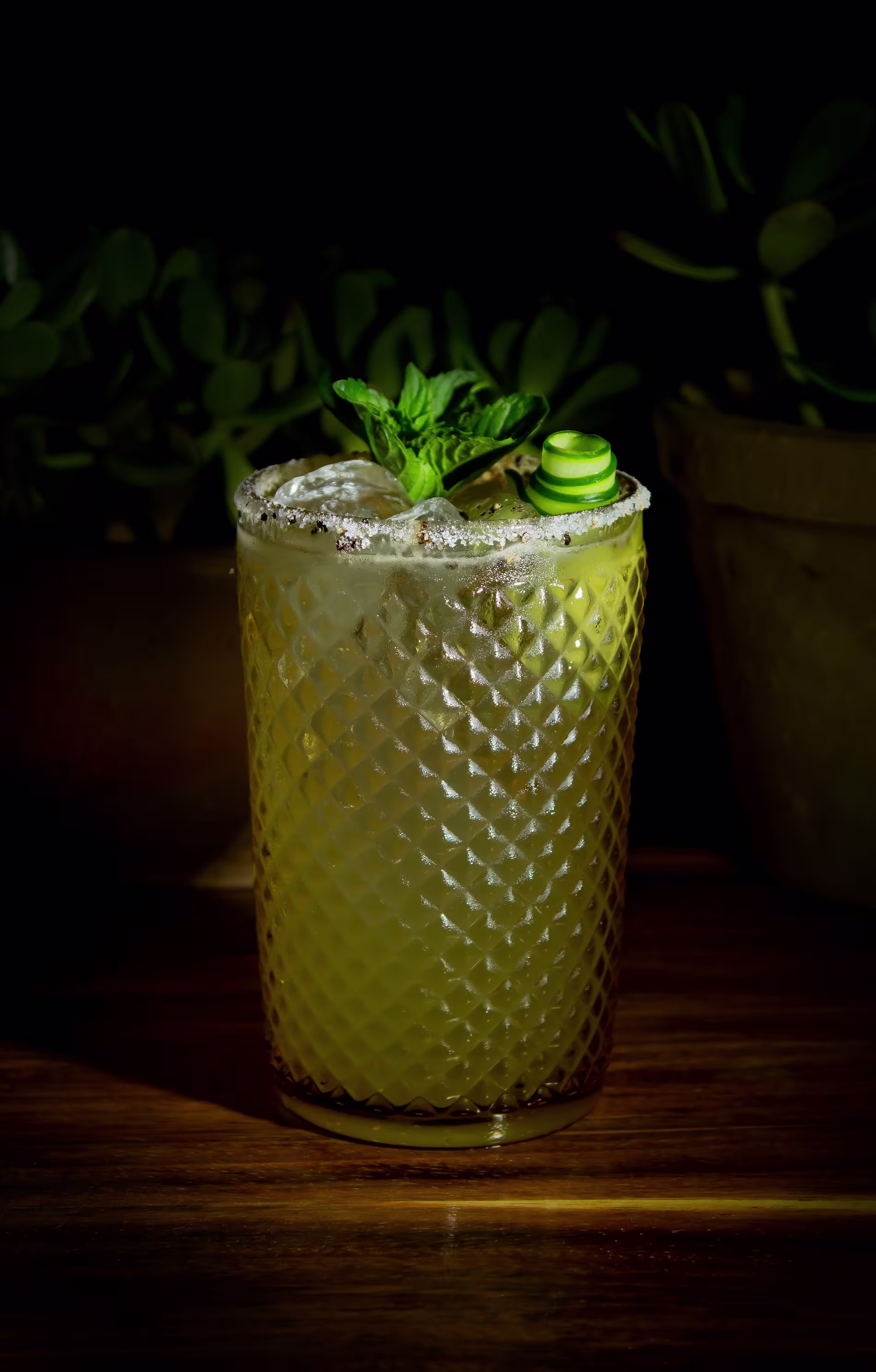 A textured glass filled with a yellow cocktail, garnished with mint, cucumber, and a salted rim, sits on a wooden surface with plants in the background.
