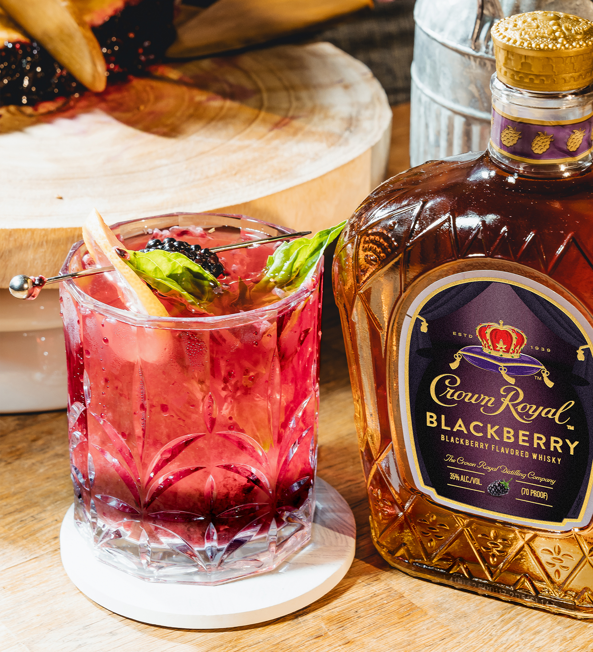 A glass of blackberry cocktail garnished with basil and fruit sits beside a bottle of Crown Royal Blackberry whisky on a wooden surface.