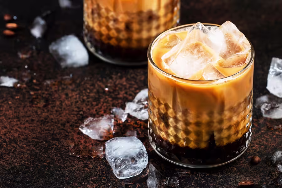 A glass of iced coffee with milk and ice cubes, placed on a dark surface with scattered ice cubes around.