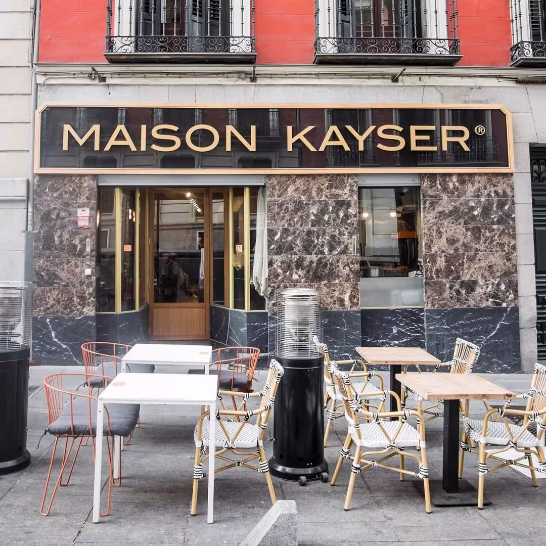 Fachada de Maison Kayser con sillas trenzadas y mesas al aire libre frente a una pared de mármol oscuro y una fachada rojiza elegante.