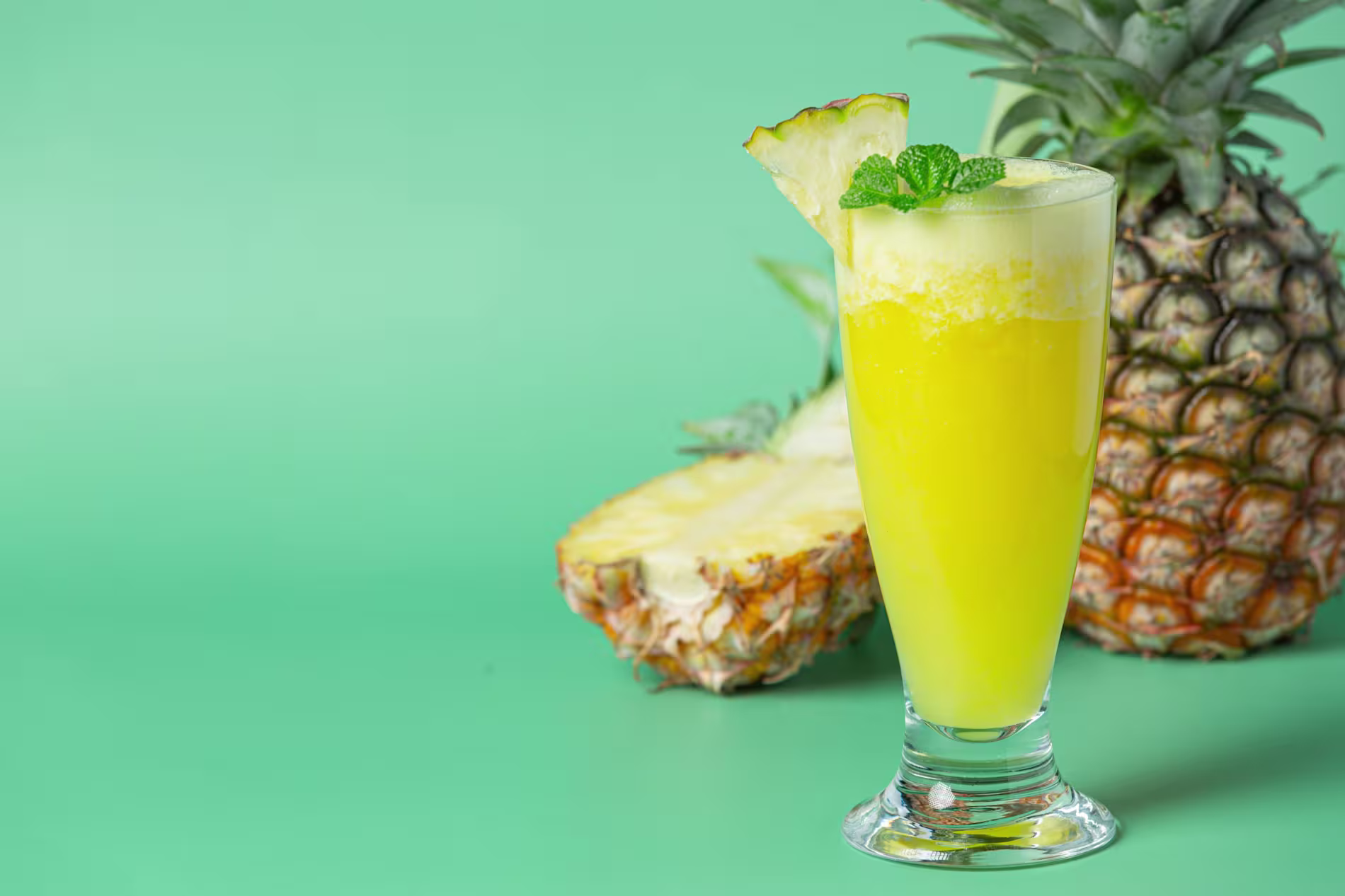 A glass of pineapple juice garnished with a pineapple slice and mint, with a whole and sliced pineapple in the background on a green surface.