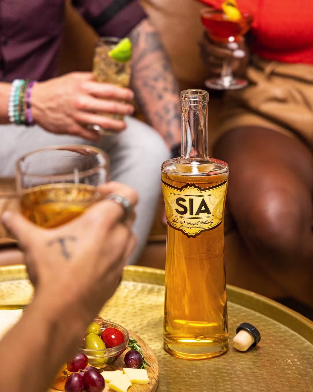 A bottle of SIA whisky on a table with a glass of whisky, a plate of fruit and cheese, and people holding drinks in the background.