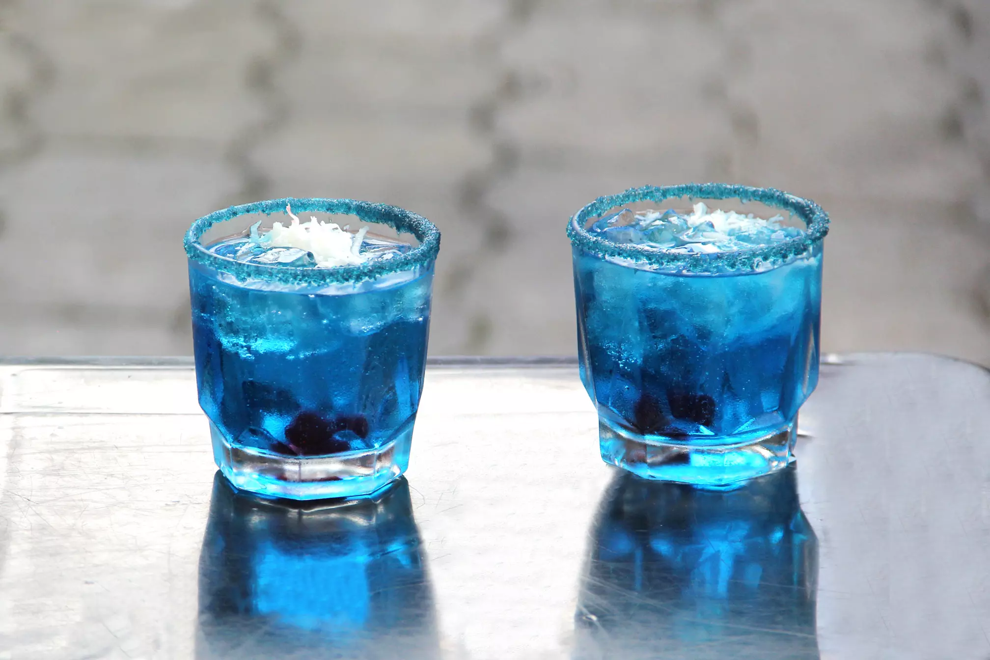 Two short glasses filled with a blue drink, ice, and topped with shredded coconut, sit on a reflective surface. The rims are coated with blue sugar crystals.