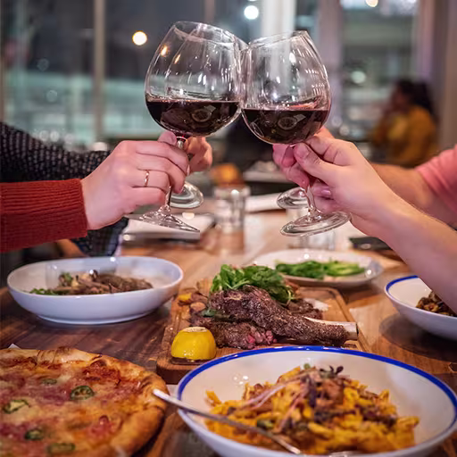 Four people toast with glasses of red wine over a table set with pizza, pasta, steak, and other dishes in a restaurant setting.