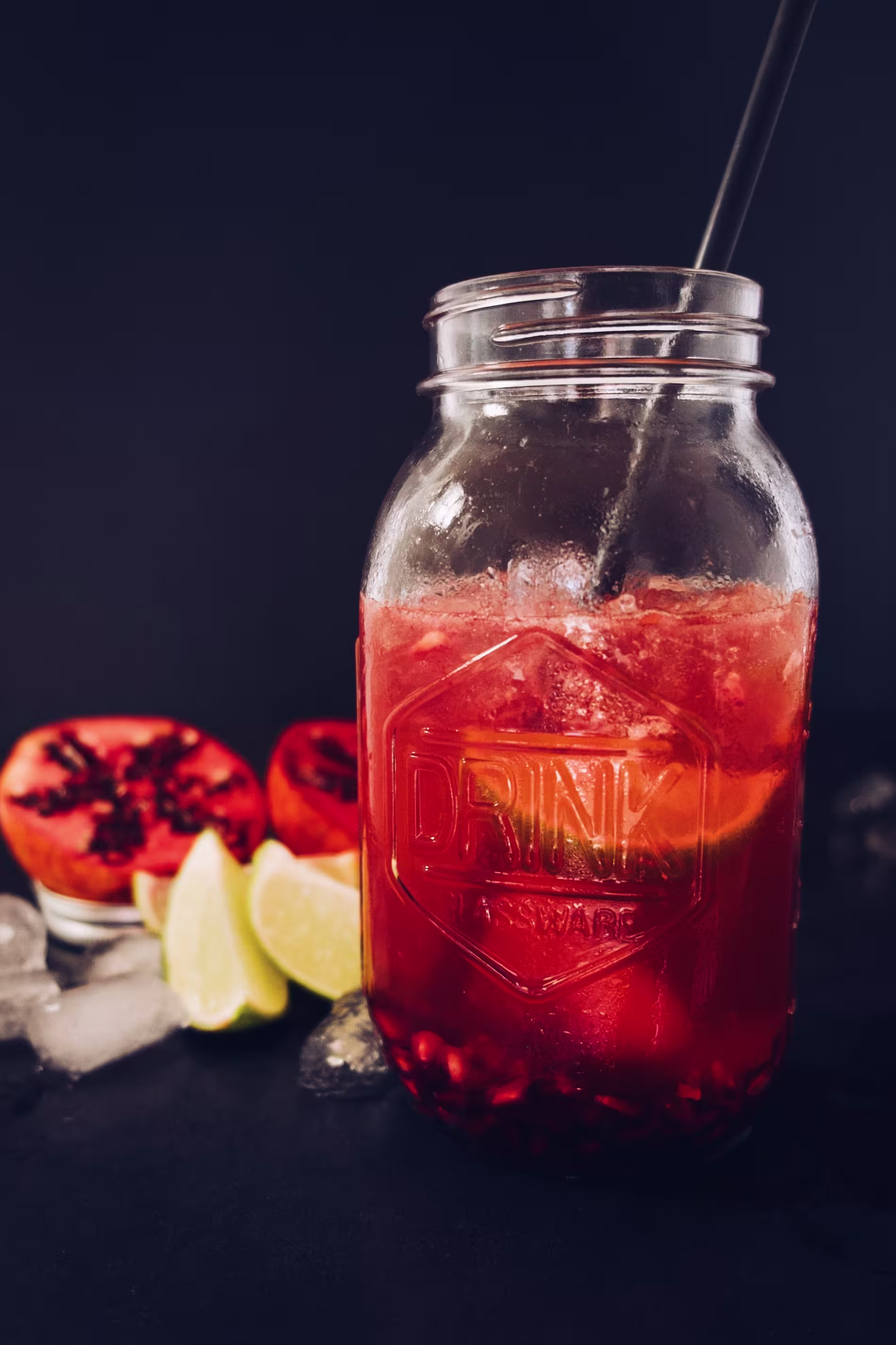 A mason jar filled with a red beverage containing ice and pomegranate seeds, with a straw, surrounded by lime wedges, pomegranate halves, and ice cubes on a dark surface.