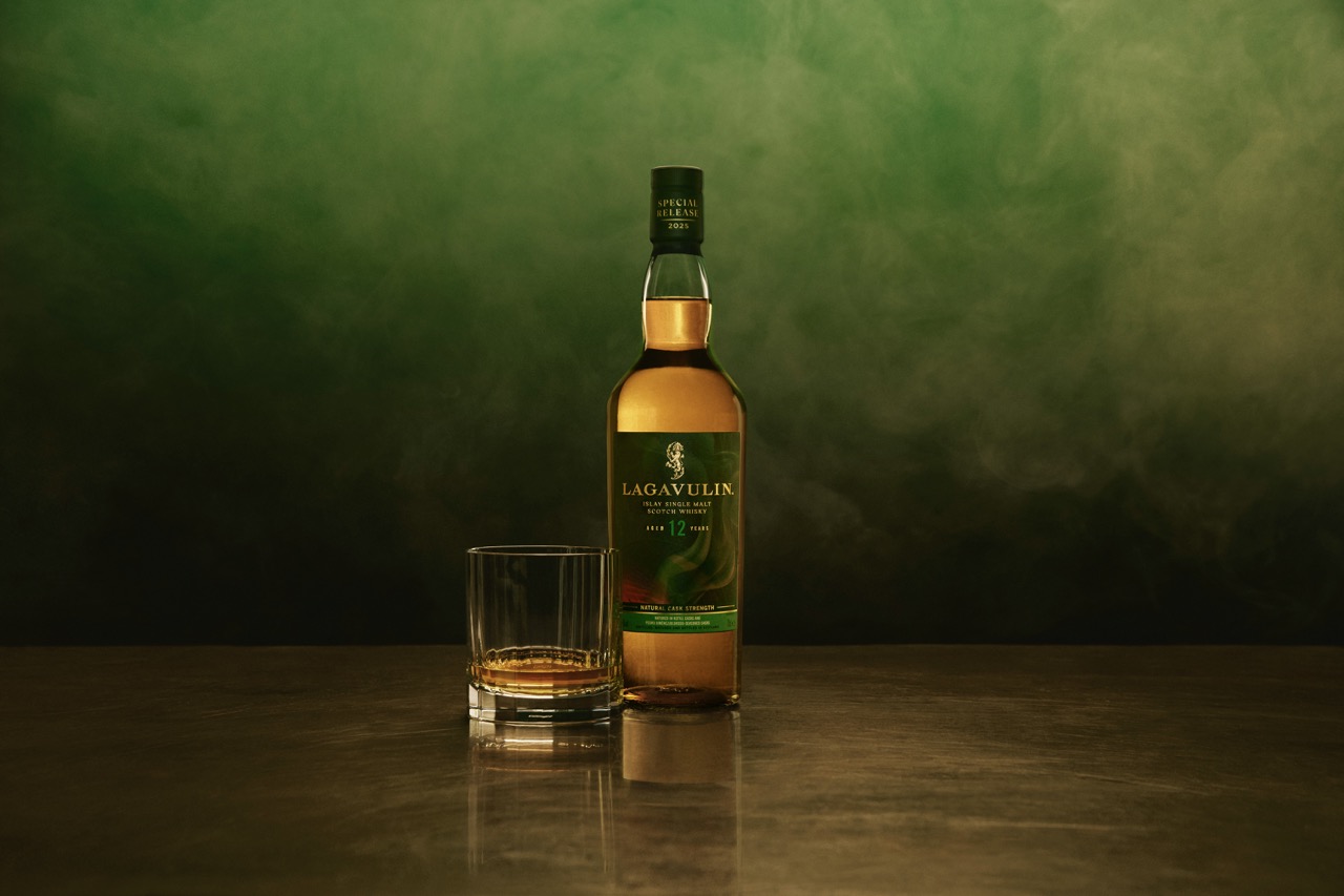 A bottle of Lagavulin 12 single malt whisky stands next to a glass with whisky, set on a reflective surface with a green smoky background.
