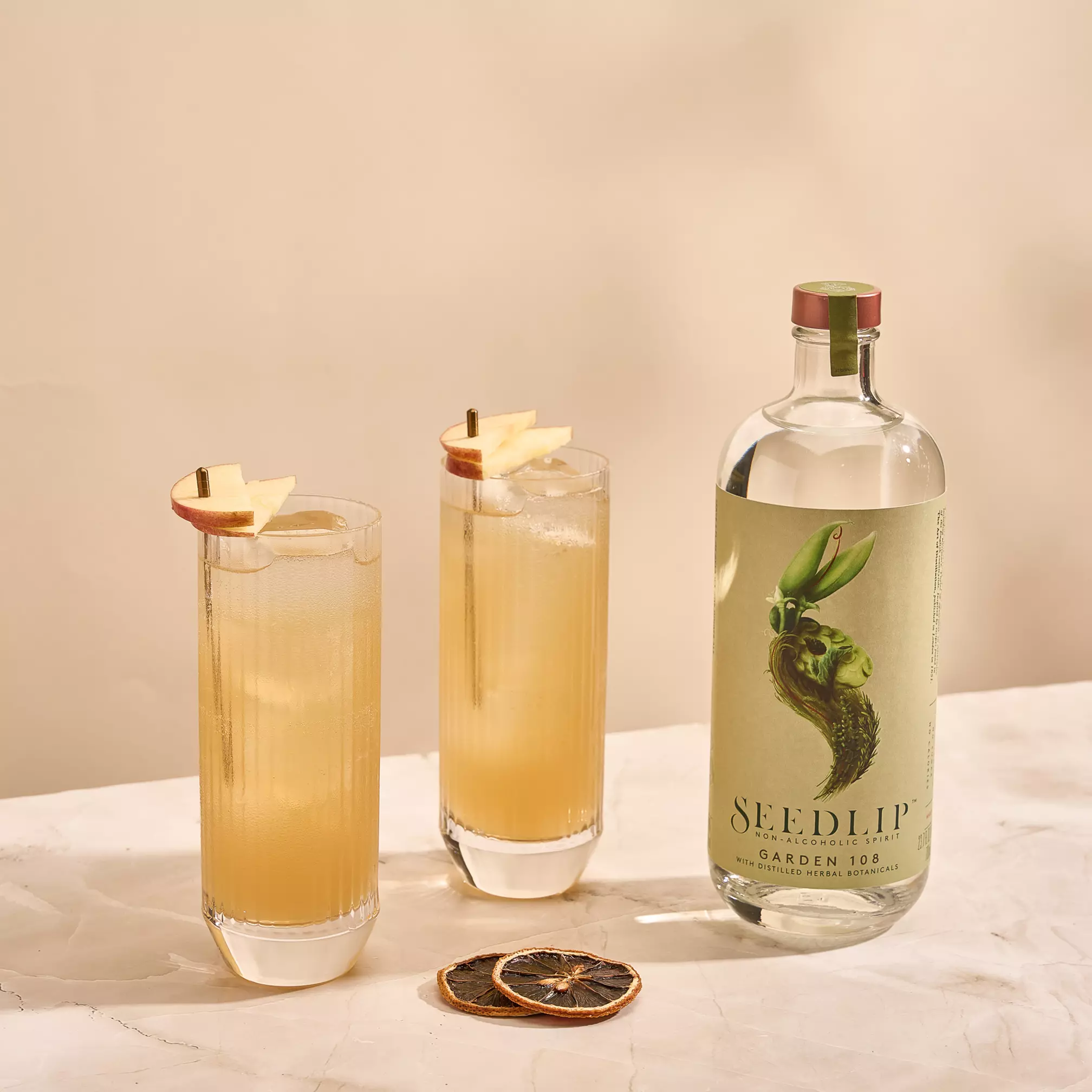 A bottle of Seedlip Garden 108 with two tall glasses of a light brown drink garnished with apple slices, and two dried citrus slices on a marble surface.