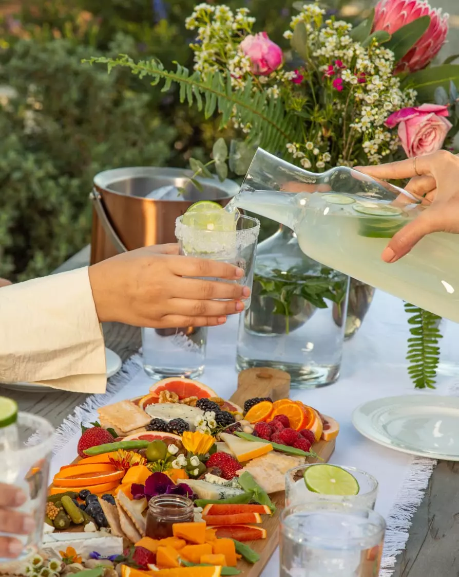 A person pours a light yellow drink from a glass pitcher into a glass with ice and lime. A charcuterie board with fruit, cheese, and bread sits on the table. Flowers are in the background.