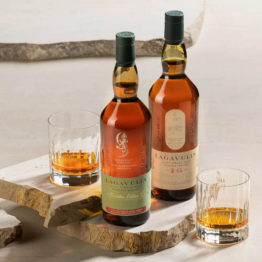 Two bottles of Lagavulin single malt Scotch whisky, each beside a glass with whisky, are displayed on stone slabs.