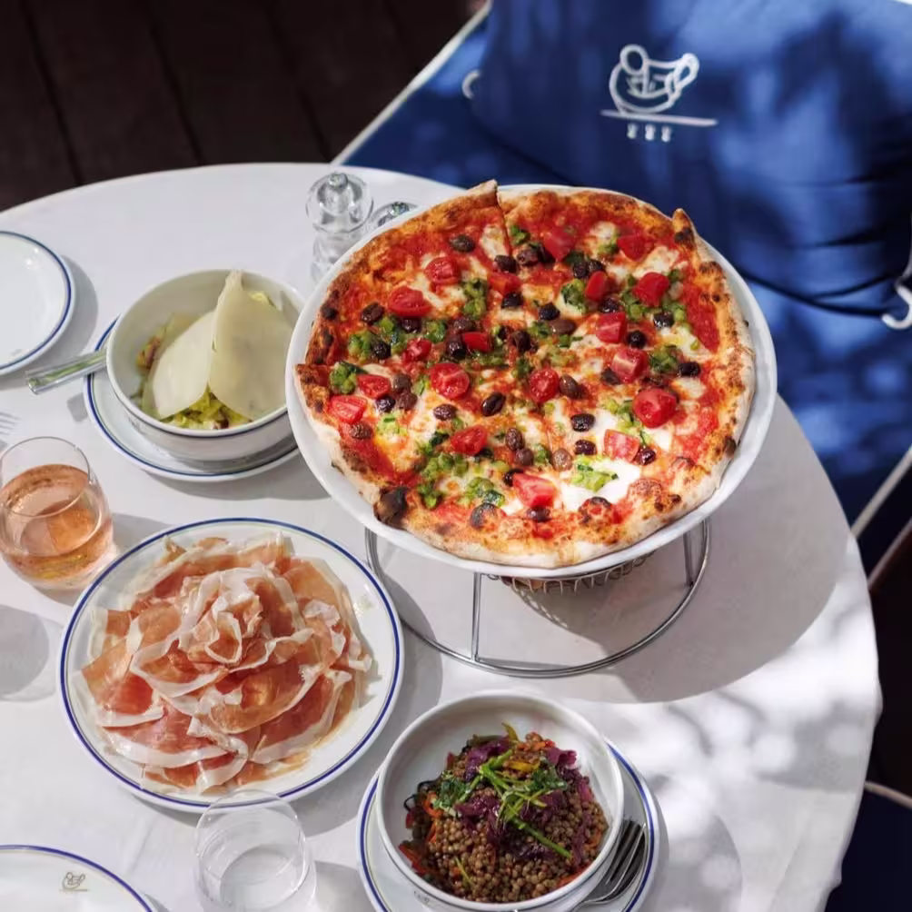 En Cipriani, una pizza fresca con tomates, aceitunas y pimientos se acompaña de jamón, ensalada y una copa de vino rosado para un almuerzo perfecto.