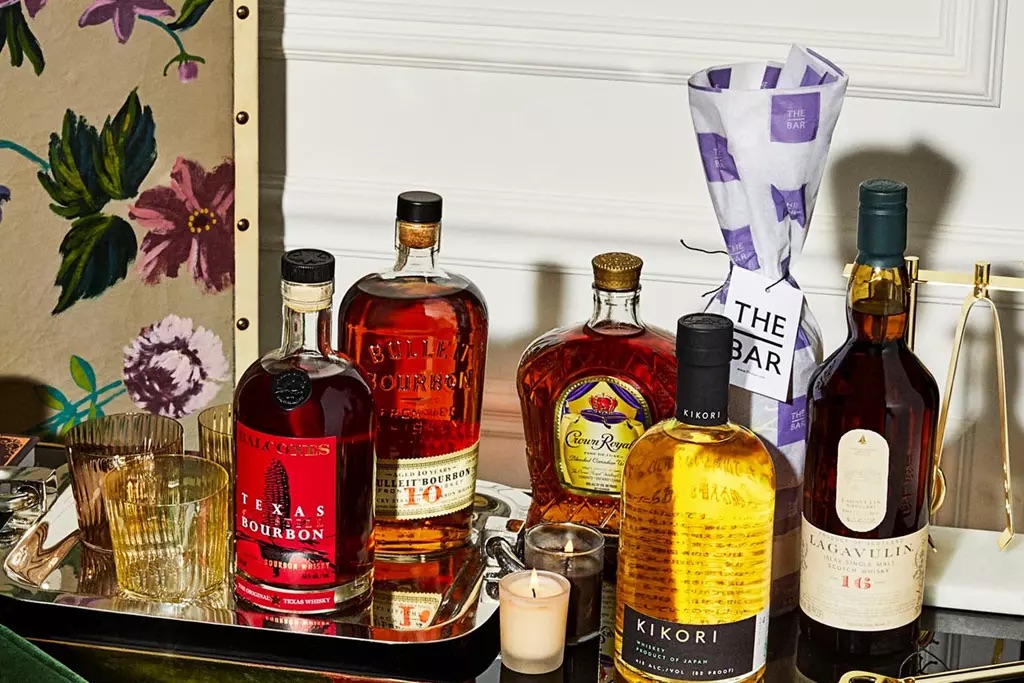 A tray holding five bottles of whiskey and bourbon, a wrapped bottle, two glasses, and a lit candle, set on a table beside a floral chair.