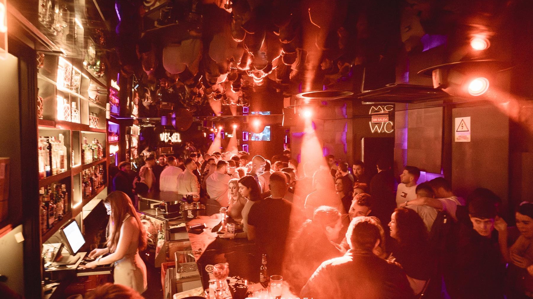 Ambiente nocturno en Vessel Club, con luces cálidas, barra llena, DJ en cabina y una pista repleta de gente disfrutando de la música y las copas.