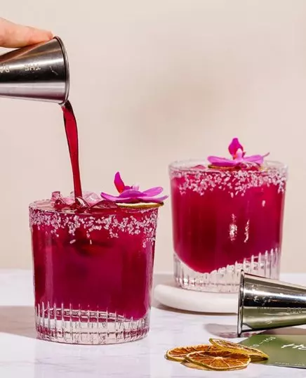 Two salt-rimmed glasses filled with a bright magenta drink, each garnished with a slice of lime and a purple flower, as red liquid is being poured into one glass.