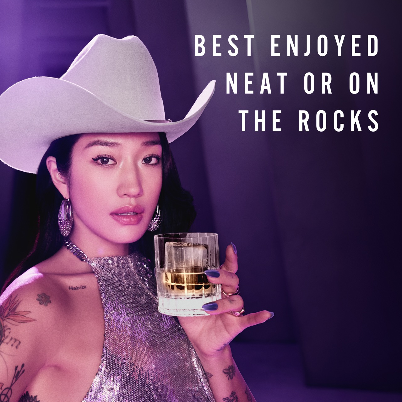 A woman in a silver halter top and white cowboy hat holds a glass of whiskey. Text reads: “Best enjoyed neat or on the rocks.” The setting is lit with purple lighting.