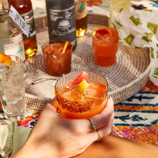 A hand holding a cocktail with chili powder on the rim and a citrus wedge, with more drinks and bottles on a woven tray in the background.