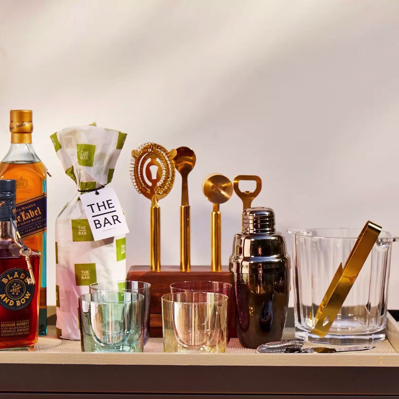 A bar setup with liquor bottles, wrapped gift, gold bar tools, cocktail shaker, ice bucket with tongs, and glassware arranged on a countertop.