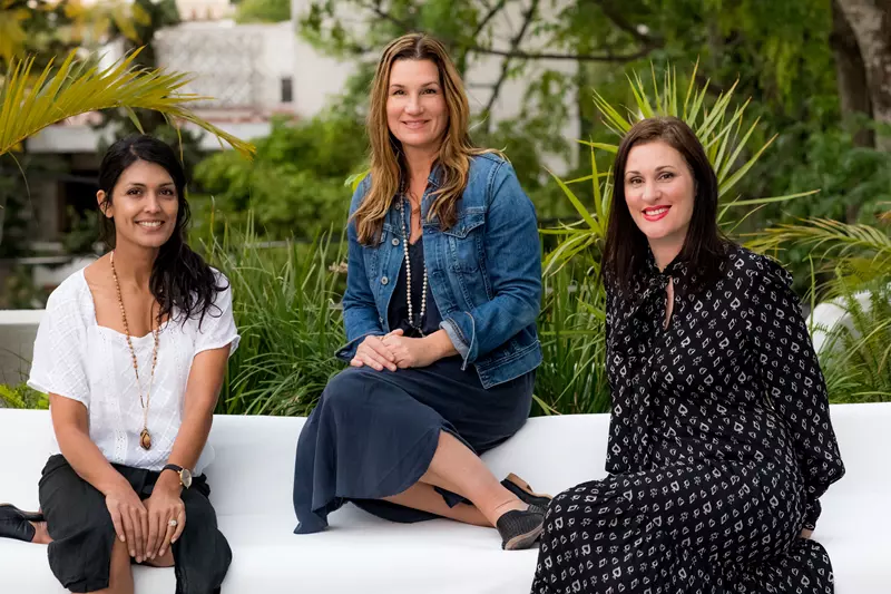 Three women sit outdoors on a white bench in front of greenery, posing for a photo. Two wear dresses, one wears a jean jacket, and one wears a white blouse with black pants.