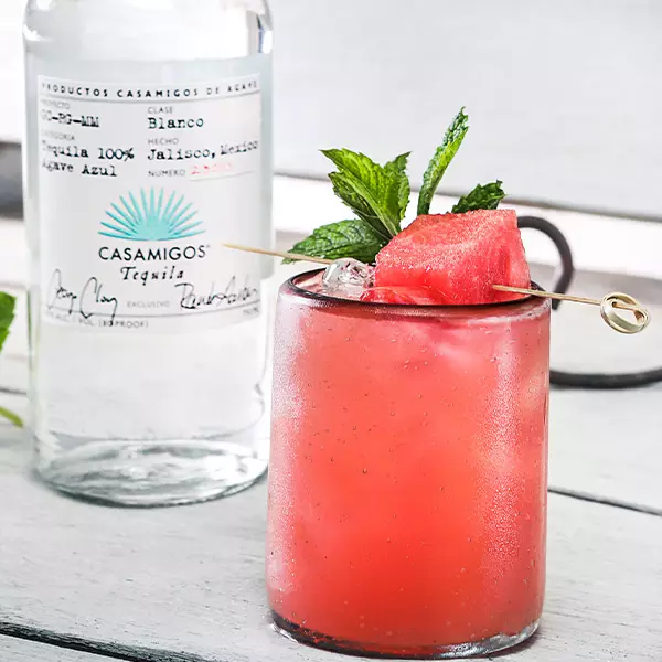 A glass of pink cocktail with ice, garnished with watermelon and mint, sits in front of a bottle of Casamigos tequila.