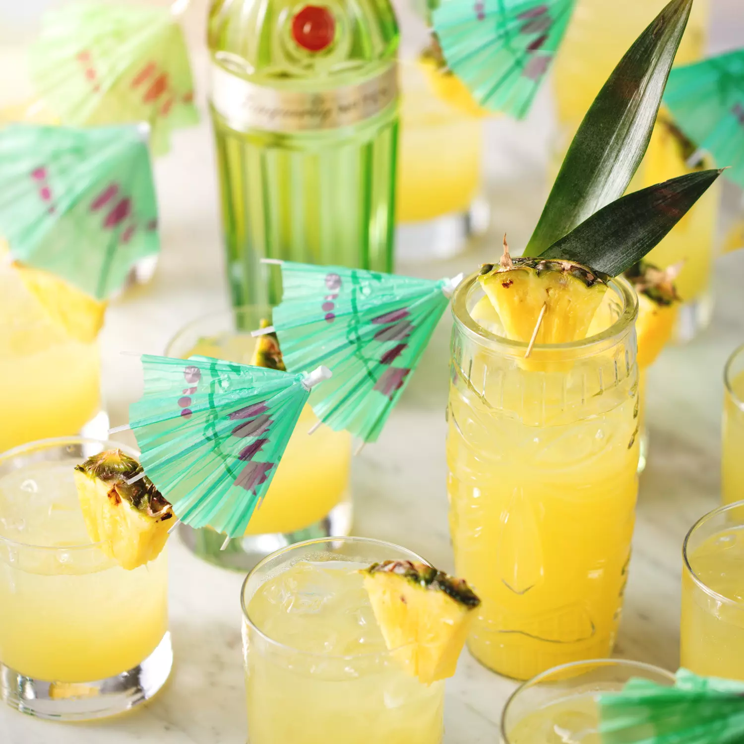 Several glasses of yellow tropical cocktails with pineapple slices and green paper umbrellas on a table, with a bottle of green liqueur in the background.
