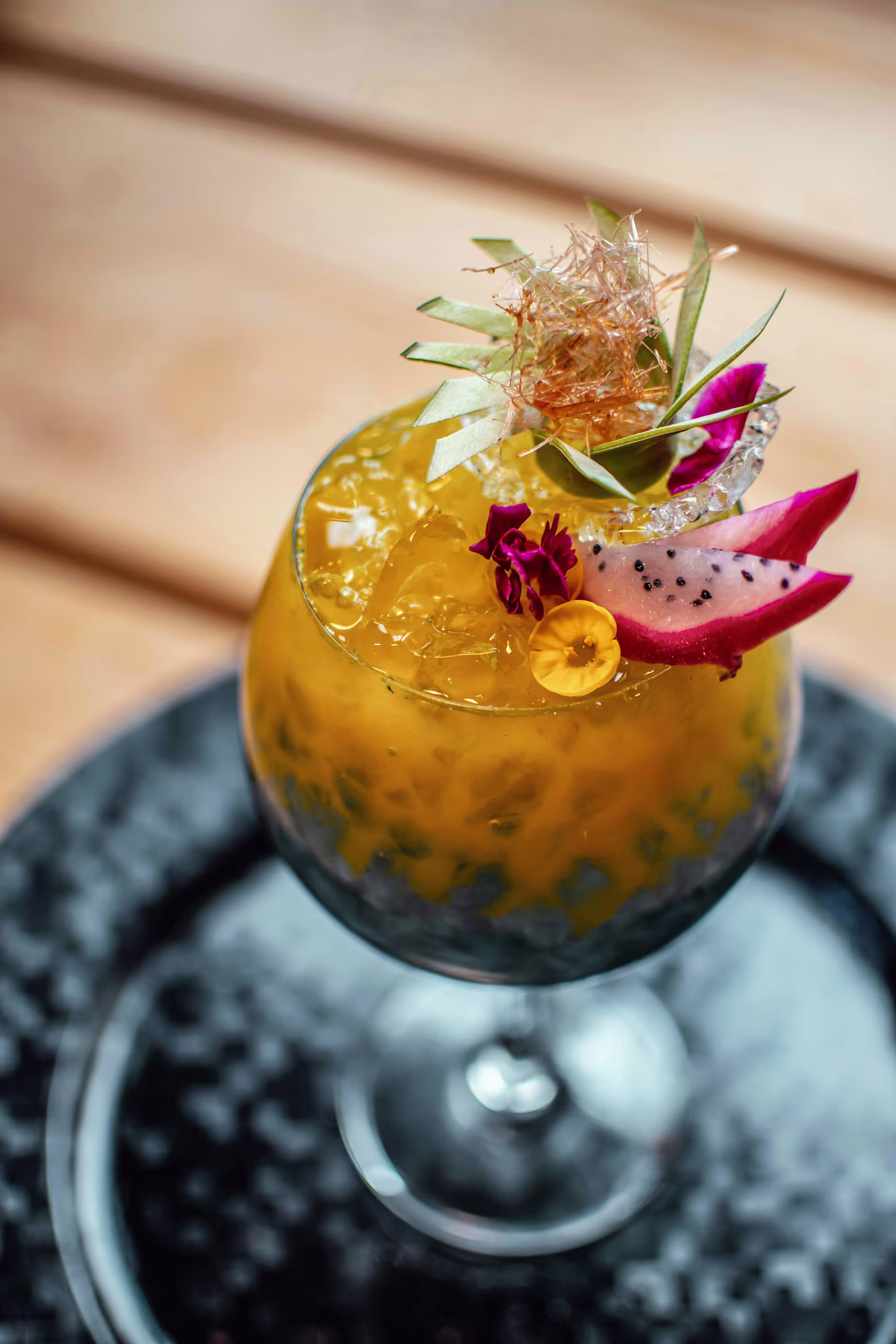 A tropical cocktail with crushed ice, garnished with dragon fruit slices, edible flowers, and a decorative sugar piece, served in a stemmed glass on a dark plate.