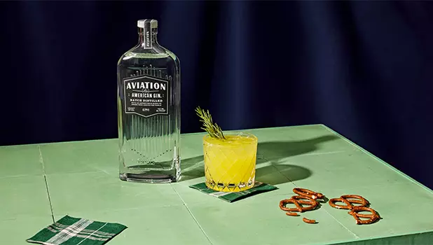 A bottle of Aviation American Gin stands next to a cocktail garnished with a rosemary sprig on a green table, accompanied by napkins and pretzels.