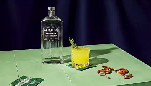A bottle of Aviation American Gin stands next to a cocktail garnished with a rosemary sprig on a green table, accompanied by napkins and pretzels.